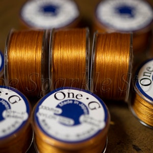 May include: Close-up of multiple spools of gold-colored thread. The spools are labeled "One.G Made in Japan 330dtex NYLON 100%".
