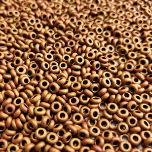 May include: A close-up image of a large quantity of small, round, matte gold beads. The beads have a hole in the center and are arranged in a random pattern.