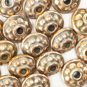 May include: A close-up of a group of small, round, copper-colored beads with a raised design. The beads have a hole in the center and are arranged in a random pattern.