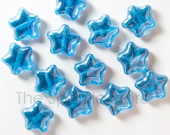 May include: A collection of translucent, blue star-shaped beads. Each bead has a swirling, marbled effect within the blue glass. The beads are scattered on a white surface, creating a celestial theme. The text "The Sparkle Store" is visible.