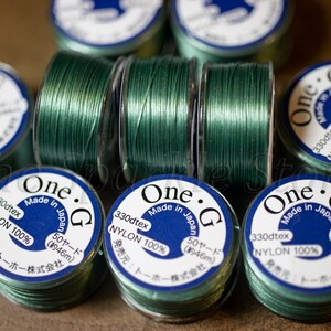 Op de afbeelding: Close-up van spoelen met groene draad met het label "One. G Made in Japan 330dtex NYLON 100% 50 yards (46m) 発売元: トーホー株式会社"