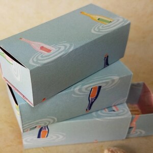 Puede incluir: Tres cajas azules apiladas con un diseño blanco y rosa de botellas de champán y ondas de agua. Las cajas son de cartón y tienen un fondo azul claro.