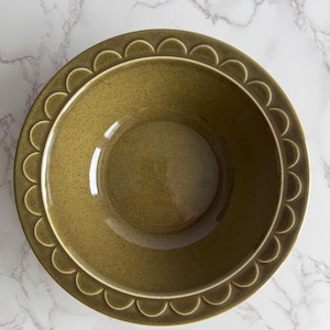 May include: Overhead view of a round, olive-green ceramic bowl. The bowl has a speckled texture and a scalloped edge design. The bowl is empty and sits on a white and gray marble surface.