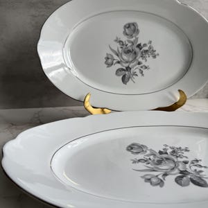 May include: Two white oval serving platters with a scalloped edge and a gray floral design. The platters have a thin gray border and a gold stand is visible behind one of the platters.