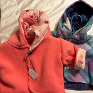 May include: Two fleece jackets on hangers. The left jacket is solid coral with a light pink floral-patterned hood lining. The right jacket features a blue and purple plaid pattern with a dark blue hood lining. Both jackets have long sleeves.