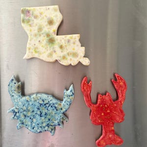 Könnte beinhalten: Drei Keramikmagnete in Form des Bundesstaates Louisiana, einer blauen Krabbe und eines roten Hummers. Der Louisiana-Magnet hat eine cremefarbene Glasur mit gelben und grünen Sprenkeln. Die Krabbe ist blau mit dunkelblauen Punkten und der Hummer ist leuchtend rot.