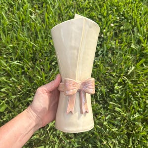 Könnte beinhalten: Eine cremefarbene Keramikvase mit glatter, glänzender Oberfläche. Die Vase hat ein gefaltetes, schriftrollenartiges Design an der Oberseite und ist mit einer geformten, hellrosa Schleife verziert, die um die Mitte des Körpers gewickelt ist.