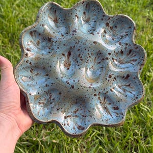 May include: A round ceramic egg serving tray with a scalloped edge and twelve circular indentations. The glaze is a light blue color with dark brown speckled patterns and feathered markings radiating from the center of each individual egg cup.