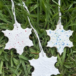 Könnte beinhalten: Drei schneeflockenförmige Ornamente in Weiß mit strukturierter Oberfläche, jeweils mit einer weißen Schnurschlaufe zum Aufhängen. Ein Ornament hat rote Sprenkel, eines blaue und gelbe Sprenkel und das dritte ist schlicht weiß und matt.