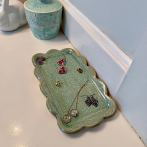 Puede incluir: Una bandeja rectangular de cerámica con bordes festoneados en un esmalte verde claro moteado. La bandeja contiene varias piezas de joyería, incluyendo dos pares de pendientes colgantes con detalles florales, un par de pequeños pendientes dorados y una cadena fina.