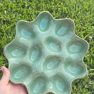 May include: A round, light green ceramic serving tray with a scalloped edge and twelve individual oval-shaped indentations. The surface features a glossy, speckled glaze finish with subtle variations in colour tone across the entire piece.