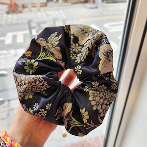 Puede incluir: Coletero de satén negro con bordado floral dorado. El accesorio para el cabello tiene forma circular, con un diseño de tela fruncida. El coletero se sostiene en una mano, con una pulsera colorida visible en la esquina inferior izquierda.