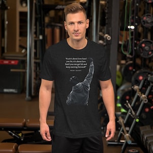 May include: Dark gray t-shirt featuring a black and white image of a boxer with his arm raised. The shirt includes a quote from Rocky Balboa: "It ain't about how hard you hit...keep moving forward!"