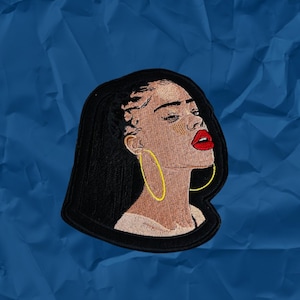 May include: Embroidered patch featuring a woman with long black hair, wearing large gold hoop earrings and red lipstick. The patch is on a blue background.