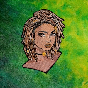 Könnte beinhalten: Gestickter Patch einer Frau mit brauner Haut und langen braunen Dreadlocks. Sie trägt eine goldene Halskette und goldene Ohrringe. Der Hintergrund ist ein grün-gelbes Batik-Muster.