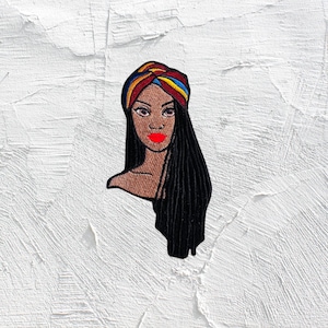 May include: Embroidered patch of a woman with long black hair, wearing a colorful head wrap. Her lips are red and she is looking to the right.