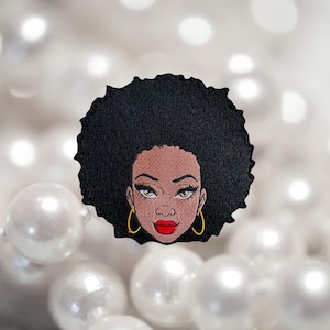 May include: Embroidered patch featuring a woman with a large afro hairstyle, brown skin, red lips, and gold hoop earrings.