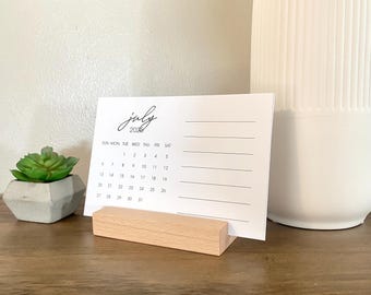 May include: A white desk calendar with a wooden stand. The calendar is for July 2025 and has the days of the week listed at the top. The calendar is on a wooden surface.