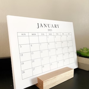 Puede incluir: Un calendario blanco con un soporte de madera. El calendario es para enero de 2025 y muestra los d&iacute;as de la semana y las fechas.