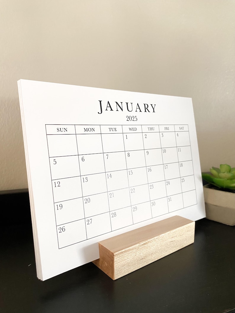 Puede incluir: Un calendario blanco con un soporte de madera. El calendario es para enero de 2025 y muestra los d&iacute;as de la semana y las fechas.