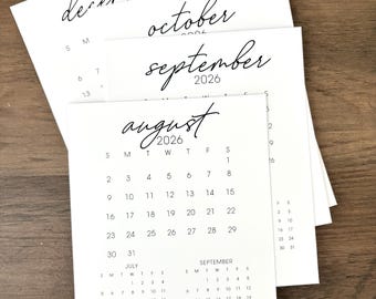 Könnte beinhalten: Ein Stapel weißer Papierkalender mit schwarzem Text. Die Monate Dezember, Oktober, September und August 2026 sind sichtbar. Die Kalender liegen auf einer Holzoberfläche.