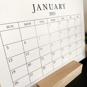 Puede incluir: Un calendario blanco con texto negro para el mes de enero de 2025. El calendario est&aacute; en un soporte de madera.
