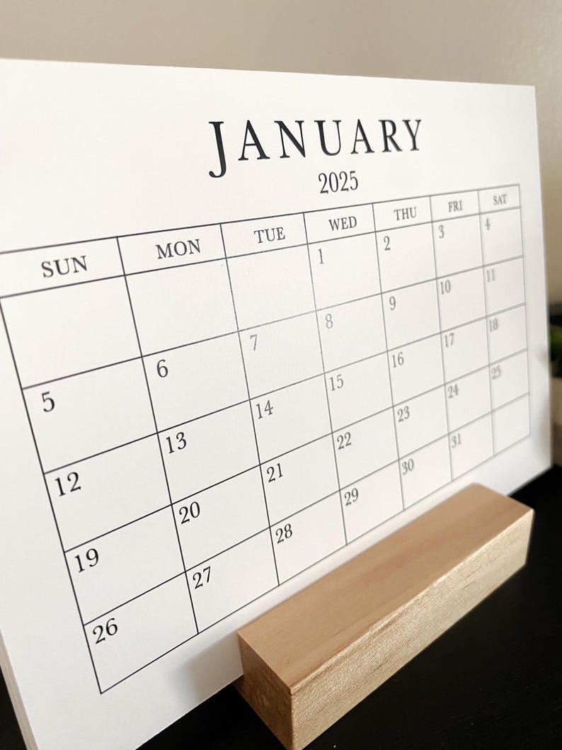 Puede incluir: Un calendario blanco con texto negro para el mes de enero de 2025. El calendario est&aacute; en un soporte de madera.