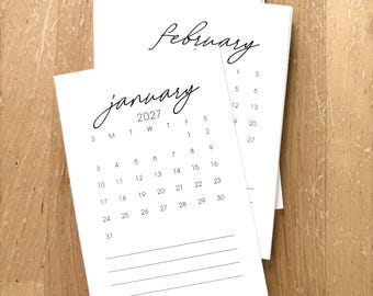 May include: A stack of white paper calendar sheets for the year 2027. The top sheet displays the month of January in a black cursive script, followed by a grid of dates and five horizontal lines at the bottom for notes. The month of February is visible.