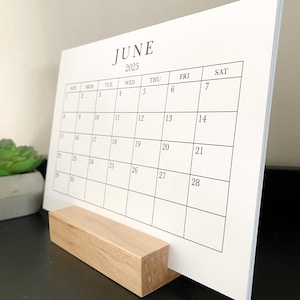 Puede incluir: Un calendario blanco con un soporte de madera. El calendario es para junio de 2025.