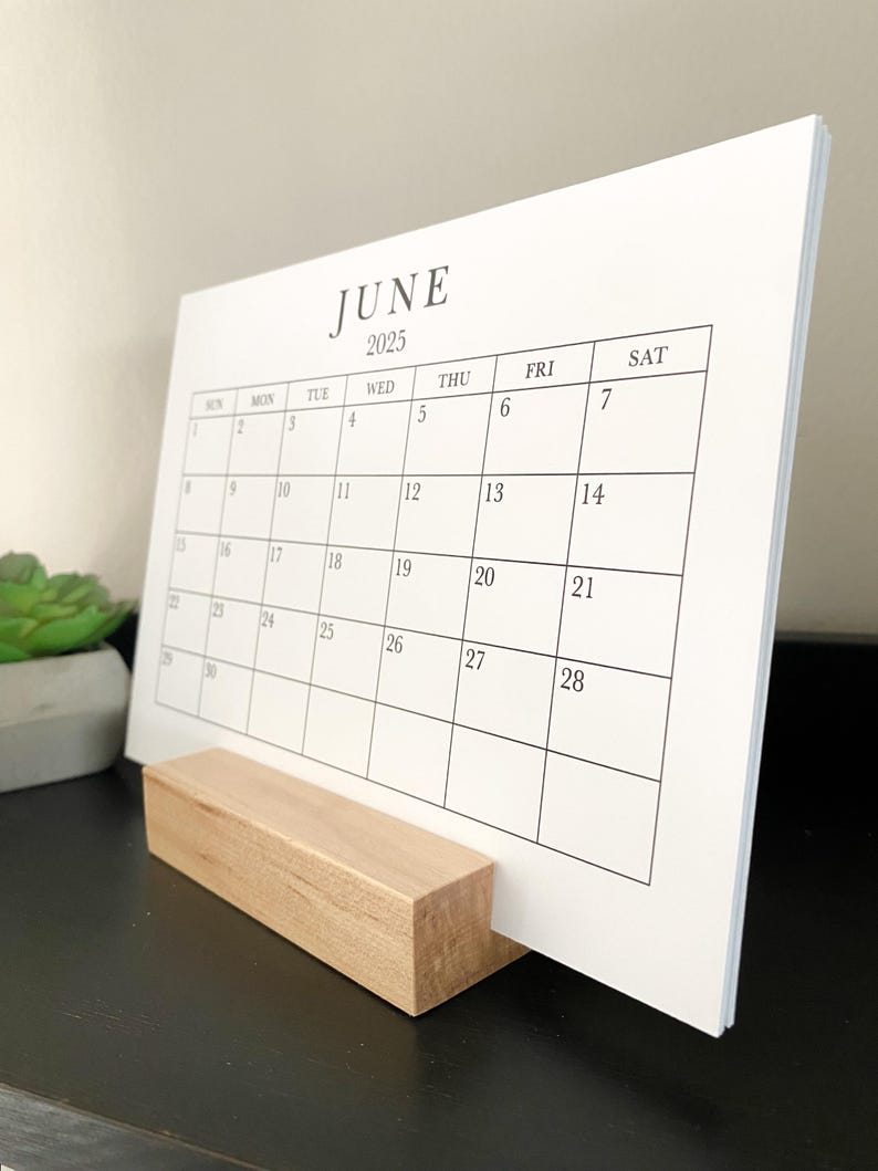 Puede incluir: Un calendario blanco con un soporte de madera. El calendario es para junio de 2025.