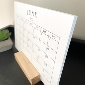Puede incluir: Un calendario de escritorio blanco que muestra el mes de junio de 2025. El calendario est&aacute; sobre un soporte de madera.
