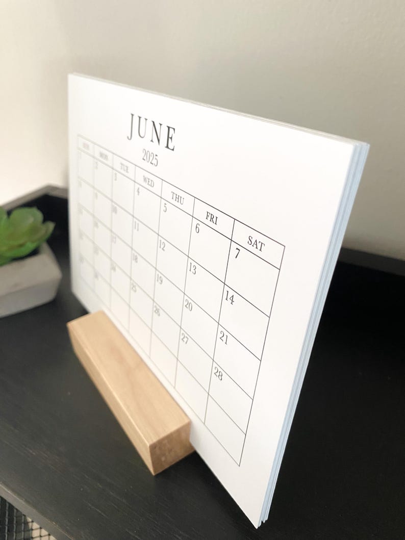 Puede incluir: Un calendario de escritorio blanco que muestra el mes de junio de 2025. El calendario est&aacute; sobre un soporte de madera.