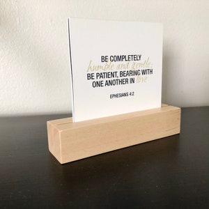 May include: A white card with black text and gold lettering on a wooden stand. The text reads "BE COMPLETELY humble and gentle: BE PATIENT, BEARING WITH ONE ANOTHER IN love EPHESIANS 4:2".