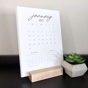 May include: A white paper calendar for January 2027 with black text, featuring a grid of dates for the month, plus smaller calendars for December and February at the bottom. The calendar is held upright by a rectangular light-colored wooden block stand.