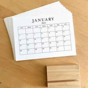 Puede incluir: Una pila de calendarios de papel blanco con un soporte de madera. El calendario es para enero de 2025.