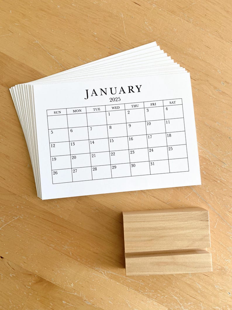 Puede incluir: Una pila de calendarios de papel blanco con un soporte de madera. El calendario es para enero de 2025.