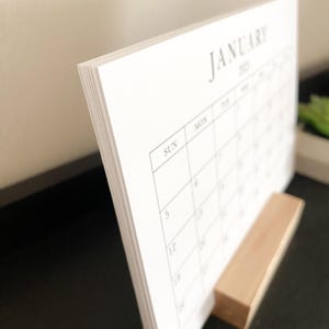 Puede incluir: Un calendario de escritorio blanco con el mes de enero de 2023. El calendario est&aacute; de pie sobre un soporte de madera.