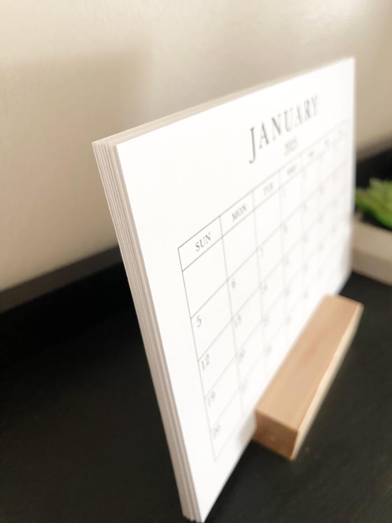 Puede incluir: Un calendario de escritorio blanco con el mes de enero de 2023. El calendario est&aacute; de pie sobre un soporte de madera.