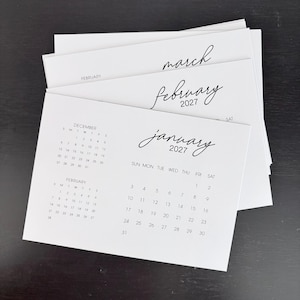 May include: A stack of white paper calendar sheets with black text. The top sheet displays the month of January 2027 with a grid of dates, while the sheets underneath show February and March 2027. The design features a clean, simple layout.