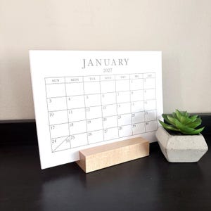 May include: A white paper calendar for January 2027 with black grid lines and text, held upright by a rectangular light-colored wooden block stand. Next to the calendar is a small green succulent plant in a gray geometric concrete planter.