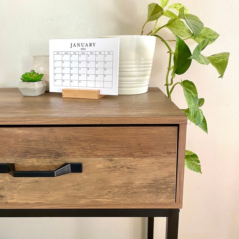 Puede incluir: Una mesita de noche de madera con un marco de metal negro y un caj&oacute;n con una manija negra. Un peque&ntilde;o calendario con el mes de enero de 2024 est&aacute; encima de la mesita de noche. Una planta en maceta con hojas verdes est&aacute; a la derecha de la mesita de noche.