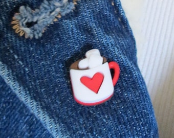May include: A red and white enamel pin shaped like a mug with a marshmallow on top and a heart inside. The pin is attached to a denim jacket.