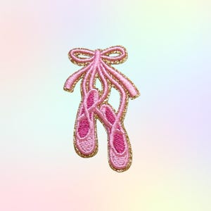May include: Embroidered patch featuring a pair of pink ballet pointe shoes with a gold glitter outline and bow. The patch is designed with a delicate, detailed stitch, perfect for adding a touch of elegance to clothing or accessories.