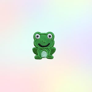 May include: A green embroidered frog patch with a smiling face and white eyes. The patch is a small, decorative item, perfect for adding to clothing or accessories. The frog is a cheerful, cartoon-like design.