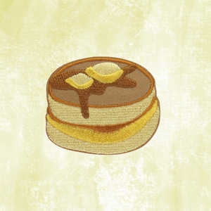 May include: Embroidered patch featuring a stack of pancakes with butter and syrup. The pancakes are brown with yellow and tan accents, and the syrup is dripping down the side. The patch is on a light yellow background.
