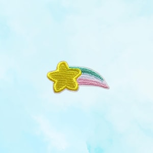 May include: Embroidered patch featuring a yellow star with a white outline and a colorful rainbow tail. The patch is set against a light blue watercolor background, creating a whimsical and decorative design.