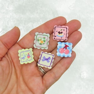 May include: A collection of six small, square, decorative patches with scalloped edges. Each patch features a different design, including a cake, cherries, a bow, and a chick. The patches are in various pastel colors.