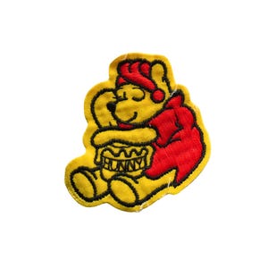 May include: Embroidered patch featuring a yellow bear wearing a red nightcap and cape, hugging a honey pot. The honey pot has the word "HUNNY" embroidered on it. The patch has a yellow border and black outlines.