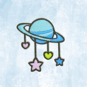 May include: Embroidered patch featuring a light blue planet with a ring, and hanging charms. The charms include pink hearts, a green heart, and blue stars. The patch is designed with a whimsical, cartoonish style.