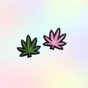 May include: Two embroidered patches in the shape of cannabis leaves. One patch is green with a black border, and the other is pink with a black border. The patches are on a pastel gradient background.